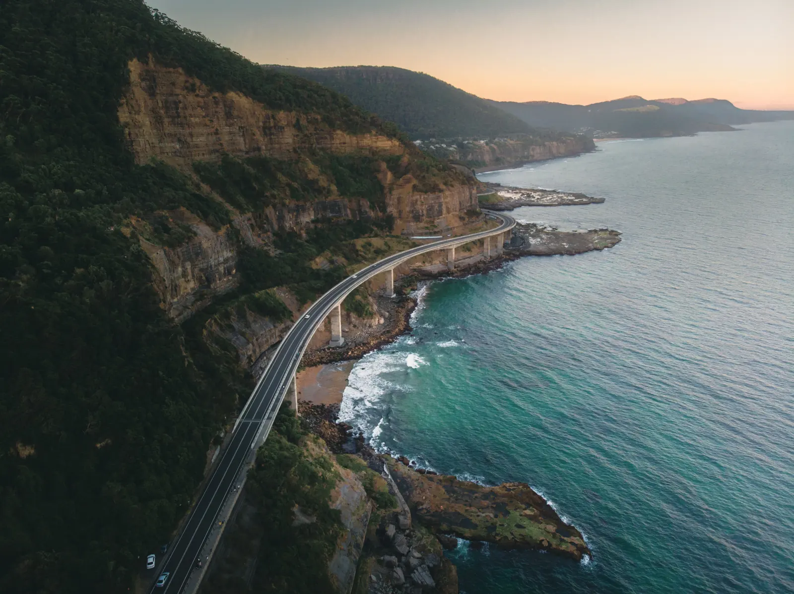 An aerial view of a steep, densely vegetated coastal escarpment meeting the sea, a road curving around the base of the cliffs on a low bridge, with the coastal strip and further headlands beyond