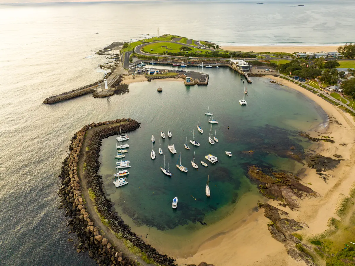 An aerial view of a harbour enclosed by two rock breakwaters, moored yachts on the water, a lighthouse on the headland beyond, and a curve of beach along one side